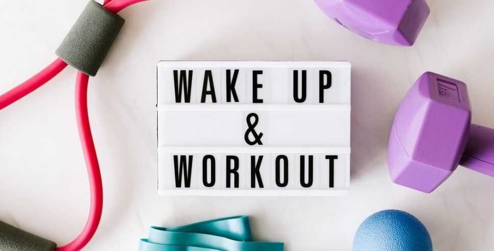 Top view closeup composition of wake up and workout inscription on light box surface among expanders and dumbbells and massage ball on white background